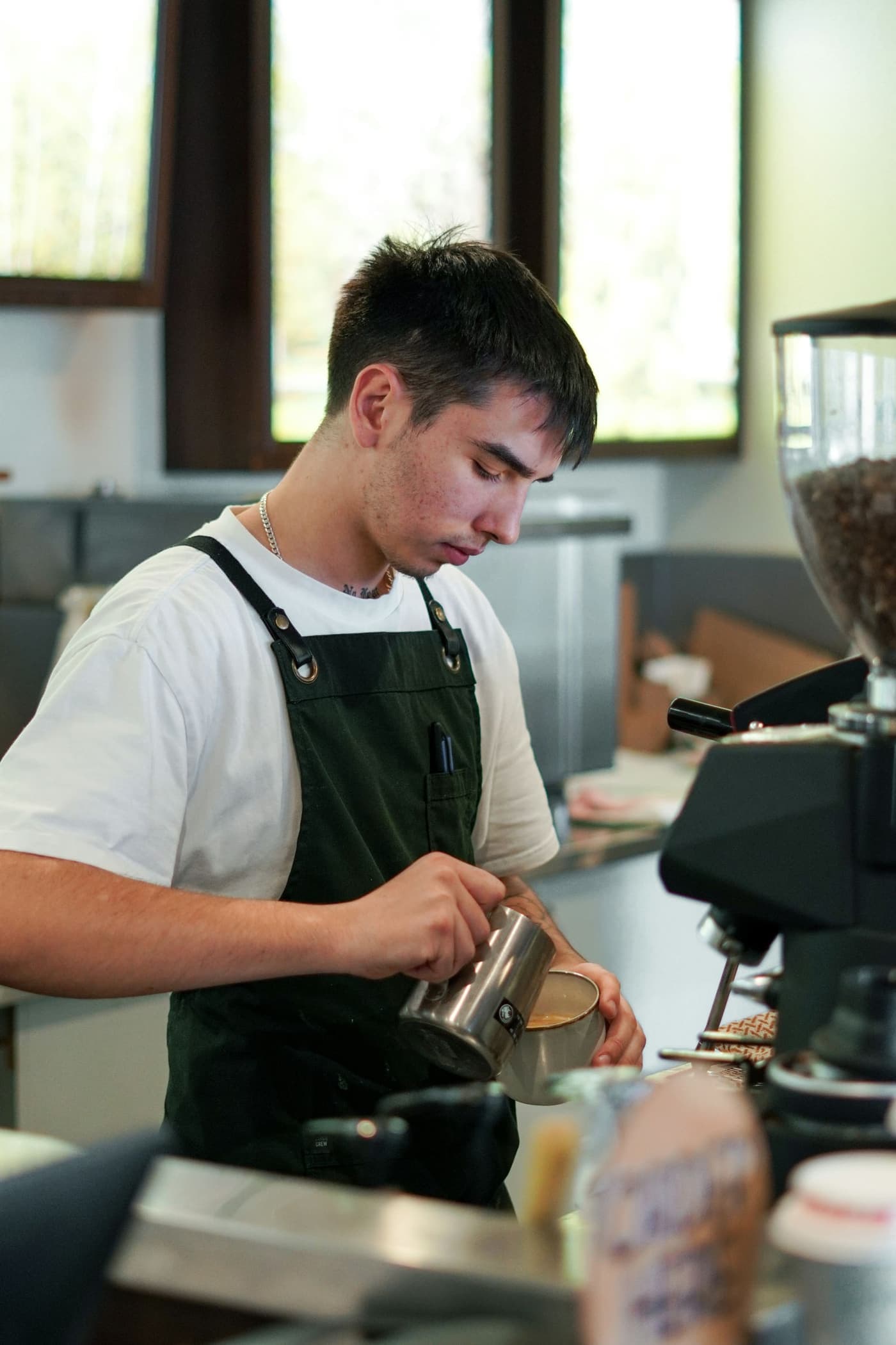 A young barista in a green apron pours steamed milk into a coffee
