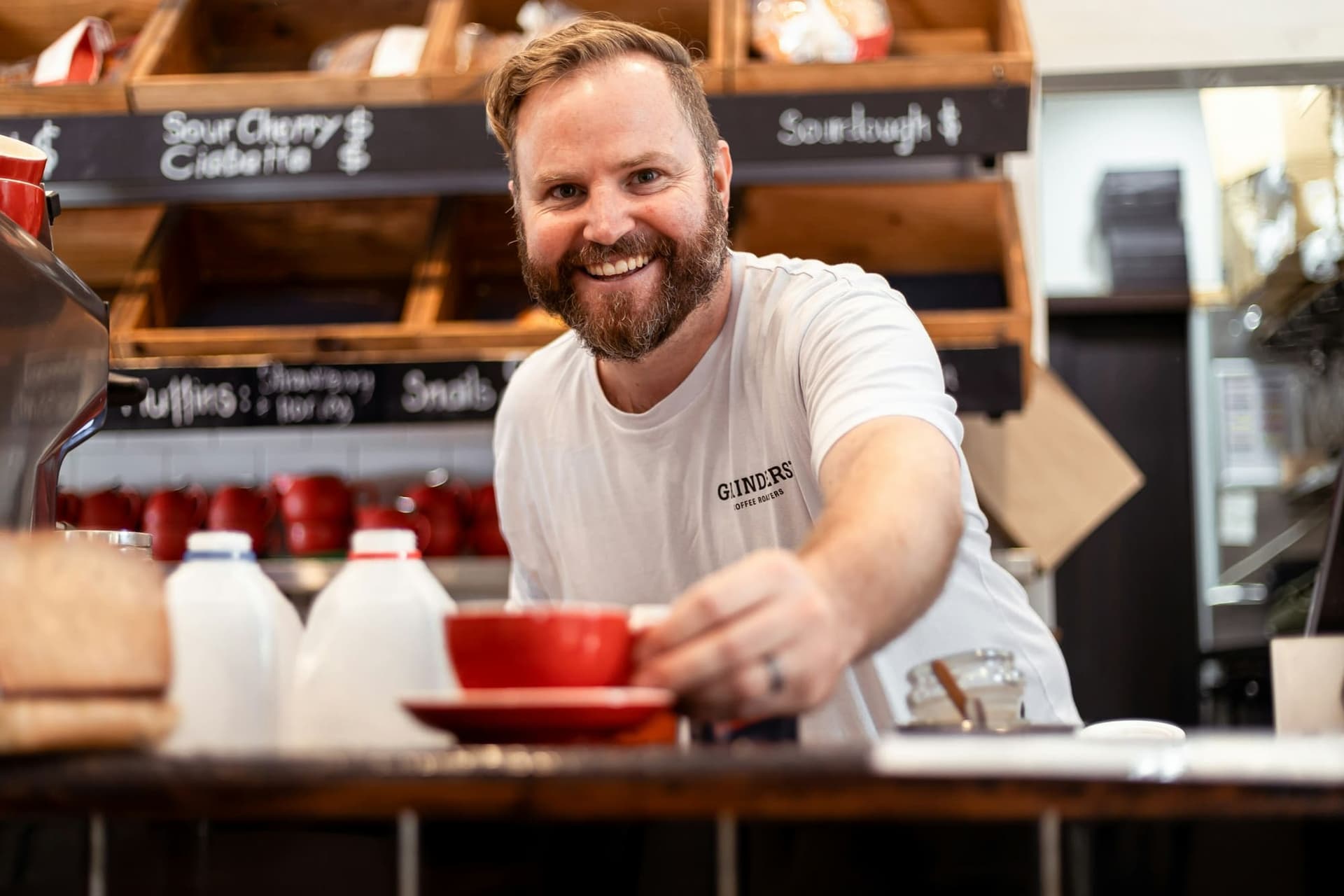 A smiling café owner hands a red cup of coffee across the counter