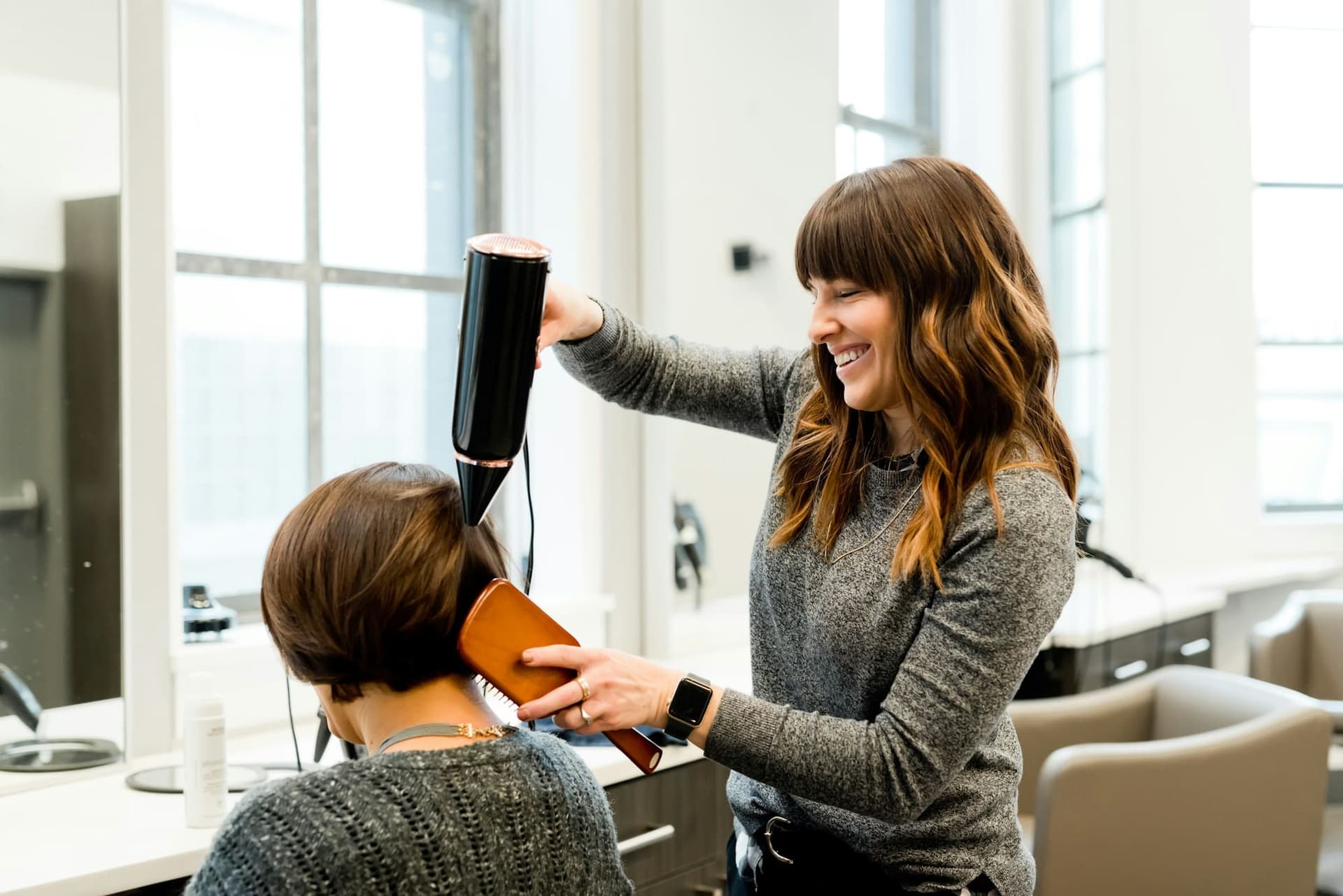 A hairdresser blow-dries a client's hair in a bright salon