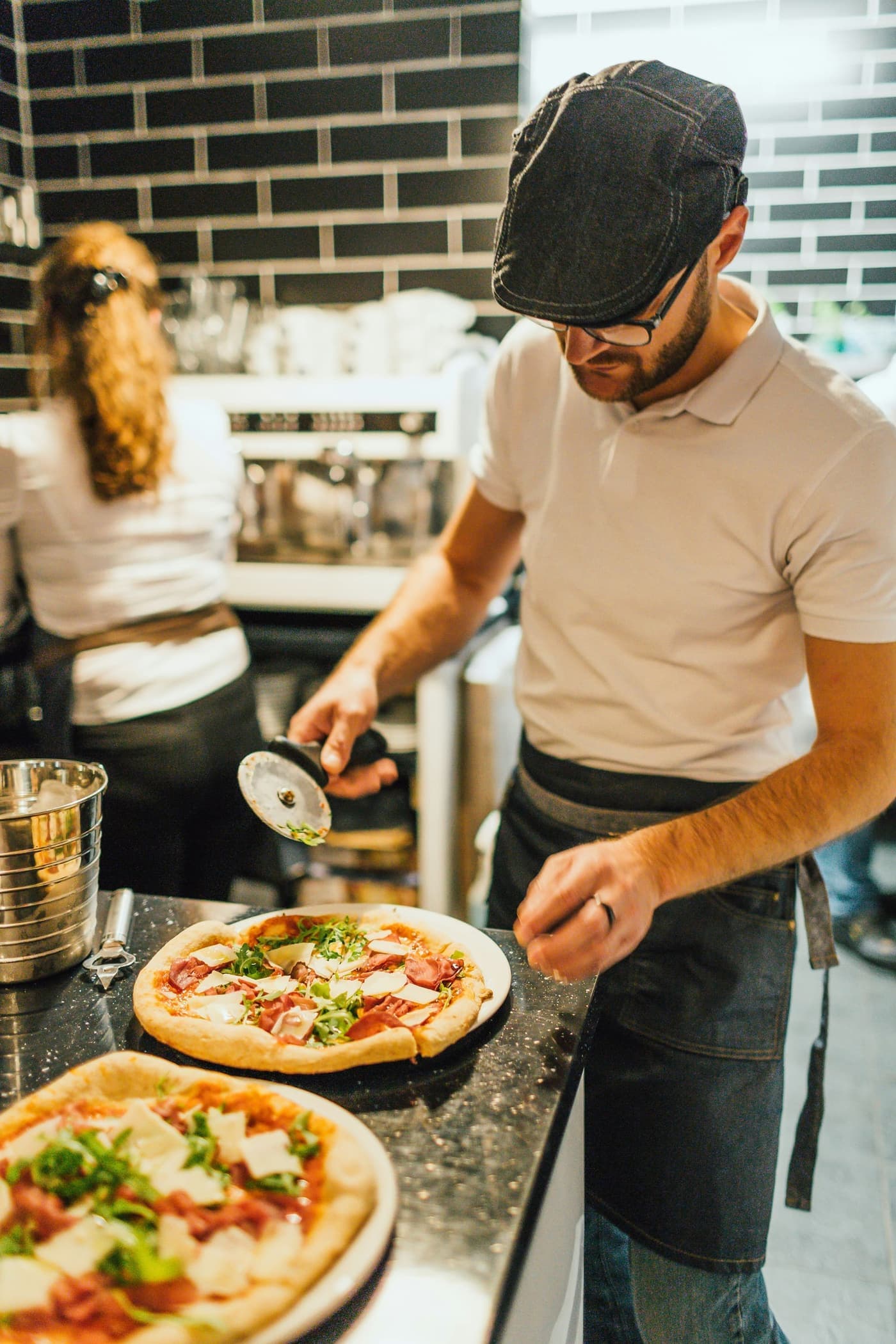 A pizza maker in a flat cap slices fresh pizzas in a small takeaway kitchen