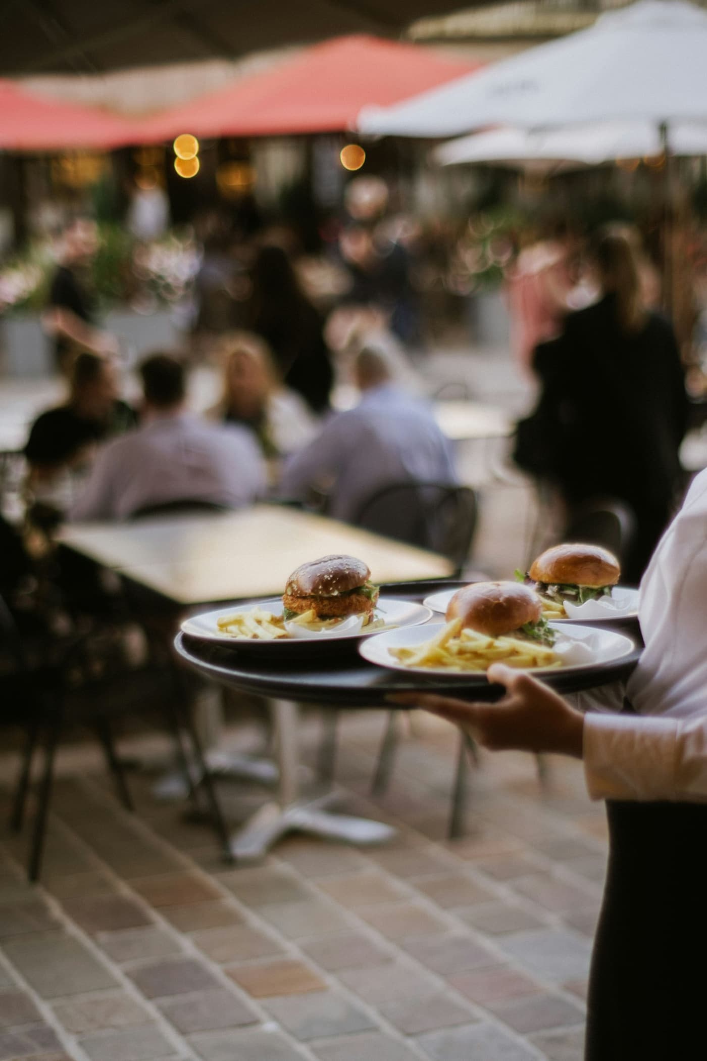 A waiter carries plates of burgers and chips across a busy restaurant terrace
